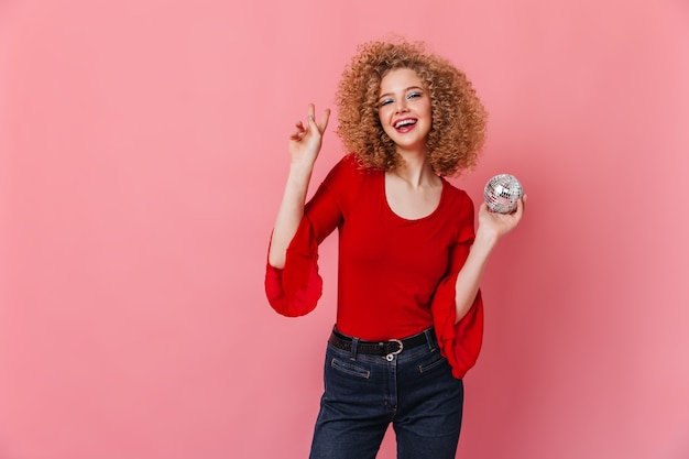 Krullend meisje met charmante glimlach toont teken van vrede. Dame in rode top met lange mouwen houdt discobal op roze ruimte.