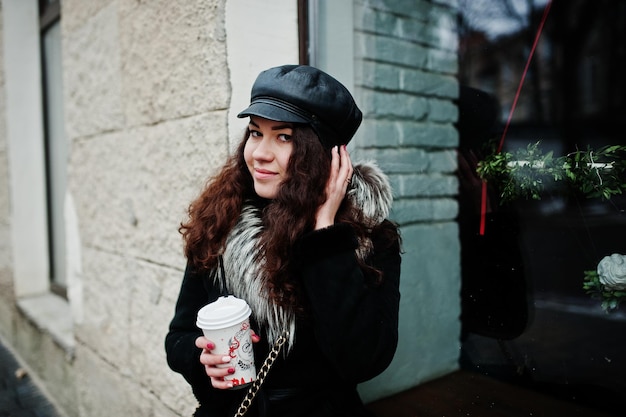 Krullend mexicaans meisje met een leren pet en een plastic kopje koffie bij de hand, wandelend door de straten van de stad