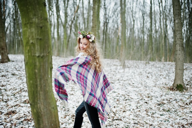 Krullend schattig blond meisje met krans in geruite plaid in het besneeuwde bos in de winterdag