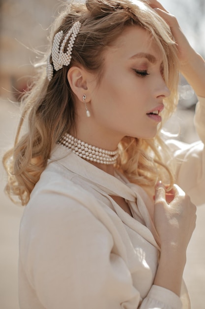 Krullende blonde jonge vrouw met pareljuwelen en gekleed in witte blouse ziet er kalm uit en poseert in het stadscentrum