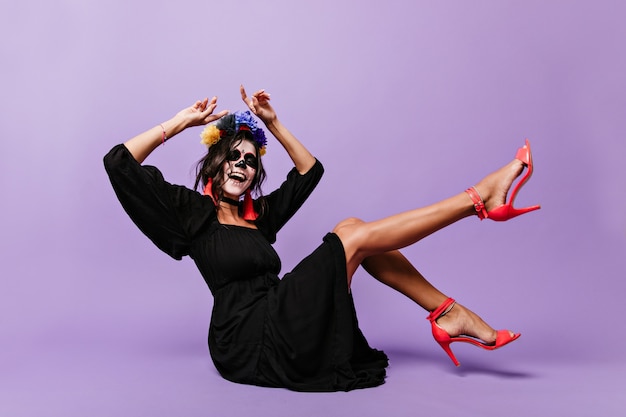 Krullende brunette met gezichtskunst voor Halloween zingt zittend op de vloer. Foto van meisje in hoge geesten op lila muur.