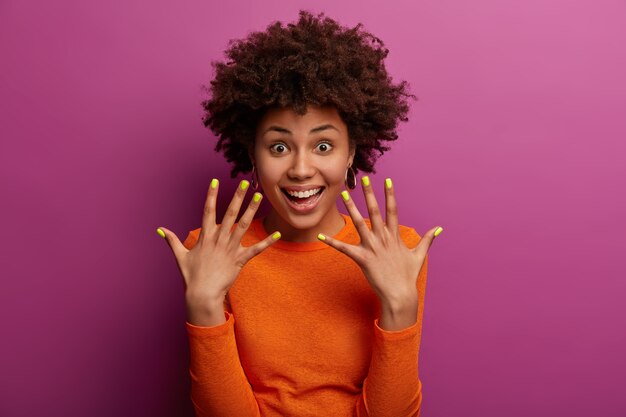 Krullende etnische vrouw toont gemanicuurde gele nagels, heeft een blije uitdrukking, lacht blij, blij na een bezoek aan de manicure, draagt een casual oranje trui, geïsoleerd over paarse muur, houdt de handen omhoog