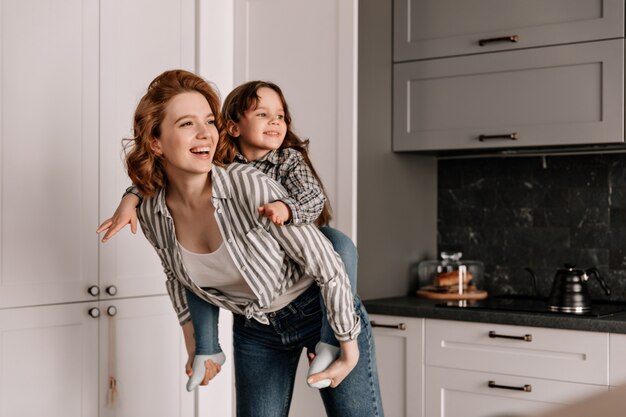 Krullende moeder in spijkerbroek speelt met haar dochter in de keuken en lacht.