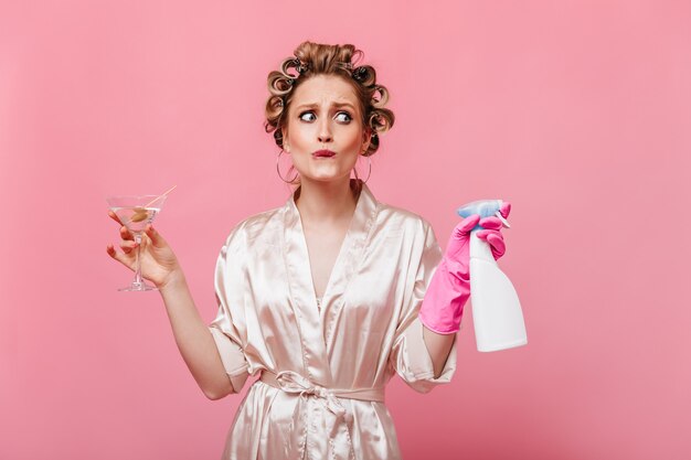 Krullende vrouw gekleed in badjas poseren op roze muur met martiniglas en wasmiddel