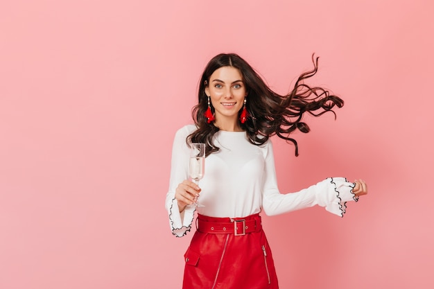 Krullende vrouw in leren rok speelt haar haren op roze achtergrond. Dame die van glas champagne geniet.