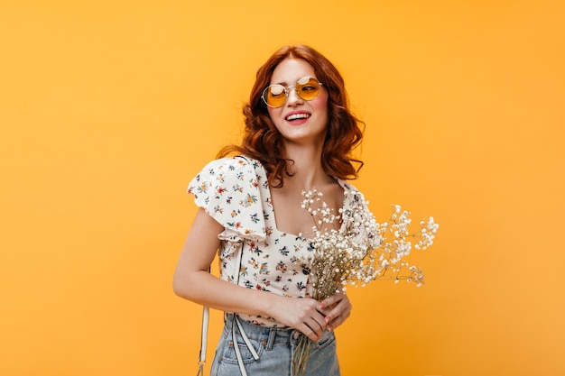 Krullende vrouw in oranje zonnebril lacht zoet en houdt wilde bloemen op oranje achtergrond.