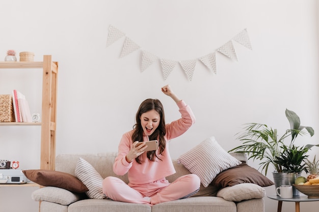 Gratis foto krullende vrouw in roze pak verheugt zich in het winnen van spel op smartphone