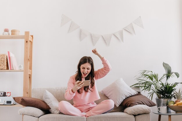 Krullende vrouw in roze pak verheugt zich in het winnen van spel op smartphone