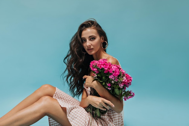 Krullende, zwartharige, mooie vrouw in zilveren oorbellen en geruite lichtroze zomerjurk die prachtige wilde bloemen vasthoudt en in de camera kijkt