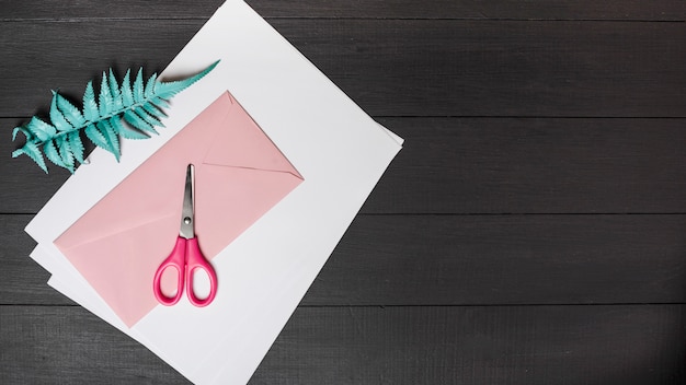 Kunstmatige varenbladeren met wit blanco papier; roze envelop en schaar op zwarte houten plank