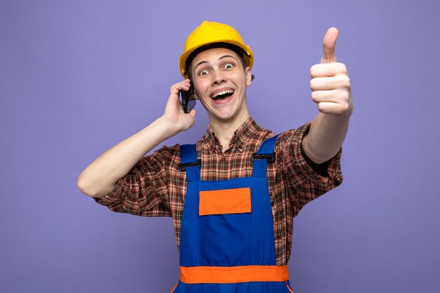 Lachen met duim omhoog jonge mannelijke bouwer die uniform draagt, spreekt op de telefoon