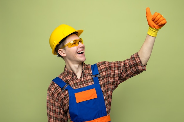 Lachen met duim omhoog jonge mannelijke bouwer met uniform en handschoenen met bril
