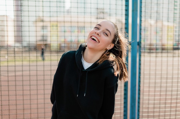 Lachen mooi meisje in donkere trui is plezier buiten op sport plein in warme lentedag close-up portret opgewonden gelukkig jonge vrouw die lacht met gesloten ogen naar zon