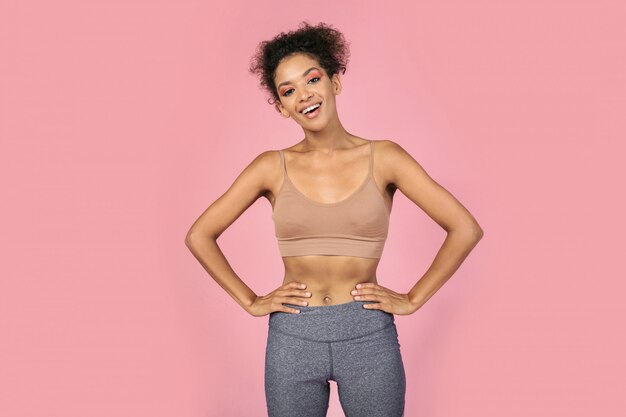 Lachen zwarte vrouw met groot figuur in stijlvolle crop top en sportieve leggins poseren op roze achtergrond.