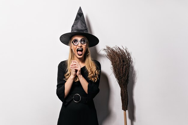 Lachend blond meisje genieten van maskerade in halloween. Goedgehumeurde heks poseren in zwarte hoed.