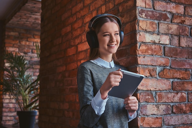 Lachend jong meisje gekleed in een elegante grijze jurk leunend tegen een bakstenen muur, luisterend naar muziek in koptelefoon en tablet.