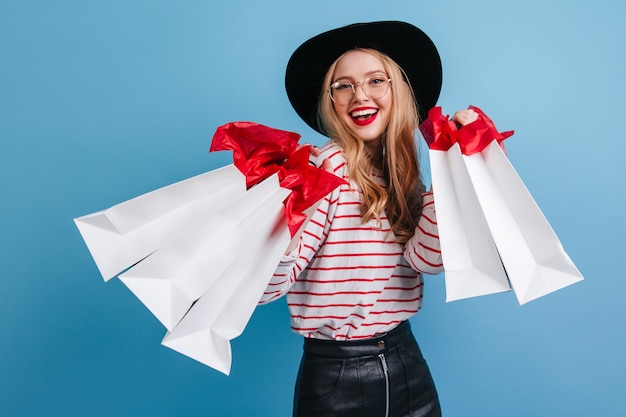 Lachend Kaukasisch meisje dat in glazen boodschappentassen houdt. Vooraanzicht van charmante shopaholic dame geïsoleerd op blauwe achtergrond.