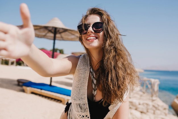 Lachend meisje in stijlvolle zonnebril met lichtbruin haar selfie maken op zee resort plezier op het strand. Portret van glimlachende jonge vrouw in gebreide kleding die voor oceaan op hemelachtergrond rust