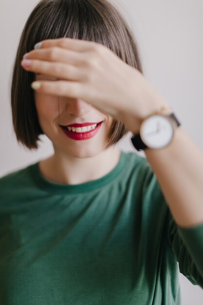 Lachend meisje met rode lippen bedekt haar ogen. Indoor foto van aangenaam vrouwelijk model in trendy polshorloge poseren.