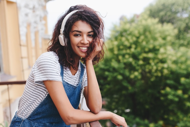 Lachend schattig meisje met trendy krullend kapsel buiten zitten en poseren op boom