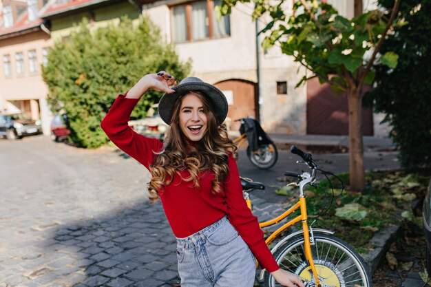 Lachend wit vrouwelijk model met glanzend krullend haar poseren in tuin met gele fiets