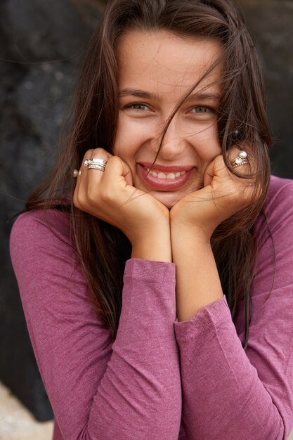 Lachende brunette blanke vrouw houdt beide handen onder de kin, drukt positieve emoties uit