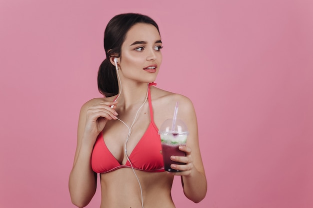 Lachende brunette in een bikini staat met oortelefoons en koude cocktail
