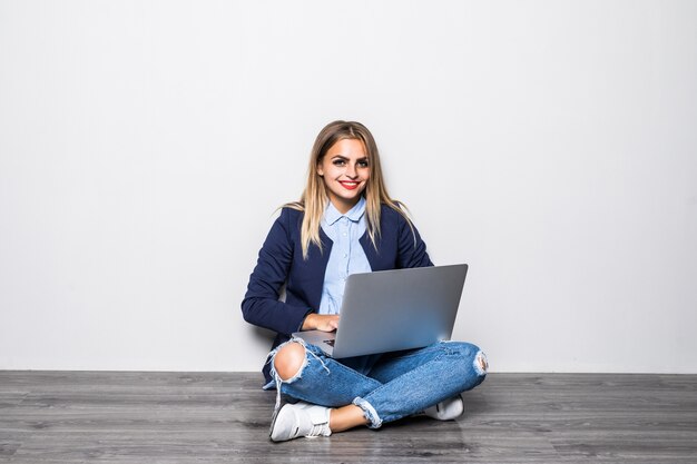 Lachende brunette vrouw in trui zittend op de vloer met laptopcomputer en wegkijken over grijze muur