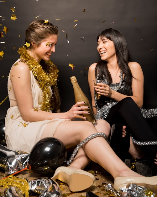 Gratis foto lachende dames in avondkleding met een glas drank en een fles op de vloer