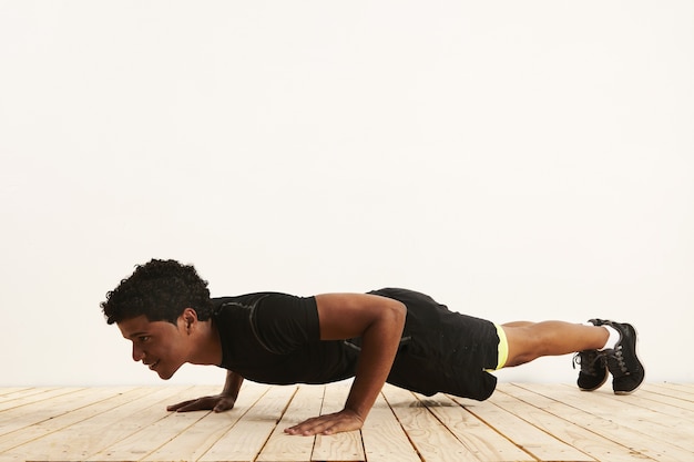 lachende fit jonge zwarte atleet doet pushups op een lichte houten vloer tegen een witte muur