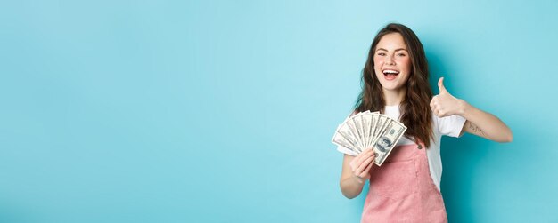 Lachende gelukkige vrouw die geld dollarbiljetten vasthoudt en duim laat zien die een snelle contante lening aanbeveelt en er tevreden uitziet terwijl ze over een blauwe achtergrond staat