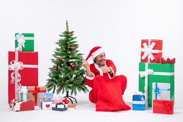 Lachende grappige jonge man verkleed als kerstman met geschenken en versierde kerstboom zittend op de grond ok gebaar maken op witte achtergrond