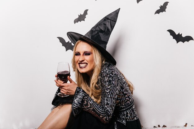 Lachende heks die bloed drinkt. Blondemeisje die in tovenaarshoed van wijn genieten bij halloween.