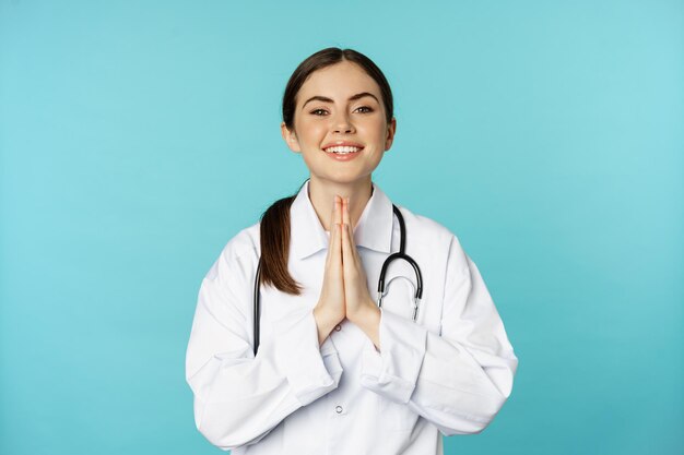 Lachende hoopvolle jonge vrouwelijke arts, hand in hand in bedelen, bedankt pose, staande over blauwe achtergrond. medisch concept