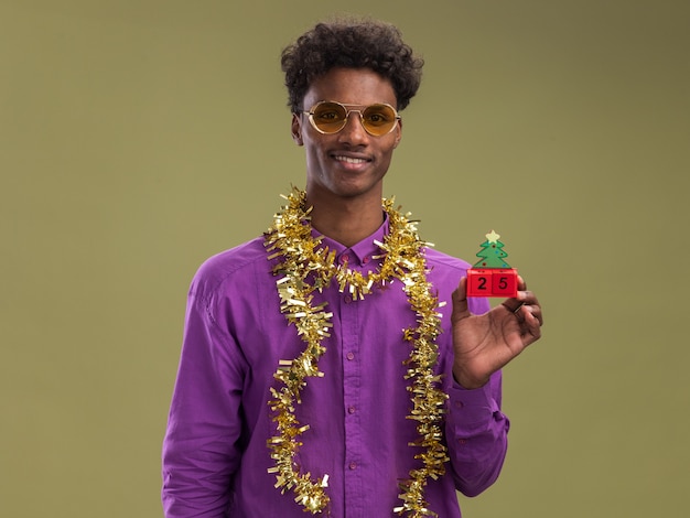 Lachende jonge Afro-Amerikaanse man met bril met klatergoud slinger rond nek met kerstboom speelgoed met datum kijken camera geïsoleerd op olijfgroene achtergrond