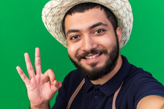 Gratis foto lachende jonge blanke mannelijke tuinman dragen tuinieren hoed gebaren ok teken en nemen selfie geïsoleerd op groene muur met kopie ruimte