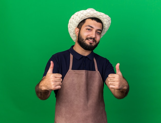 lachende jonge blanke mannelijke tuinman tuinieren hoed dragen thumbs up van twee handen geïsoleerd op groene muur met kopie ruimte