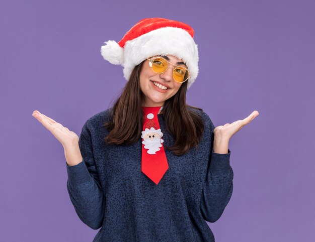 lachende jonge blanke meisje in zonnebril met kerstmuts en kerst stropdas hand in hand open geïsoleerd op paarse achtergrond met kopie ruimte