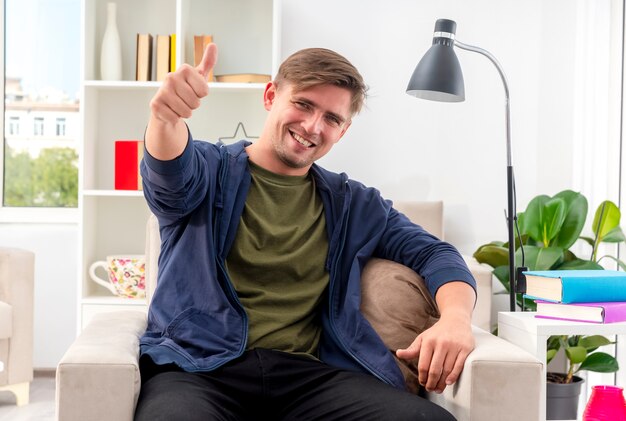 Lachende jonge blonde knappe man zit op fauteuil duimen omhoog kijken