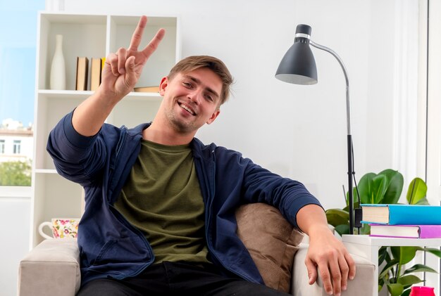 Lachende jonge blonde knappe man zit op fauteuil in de woonkamer