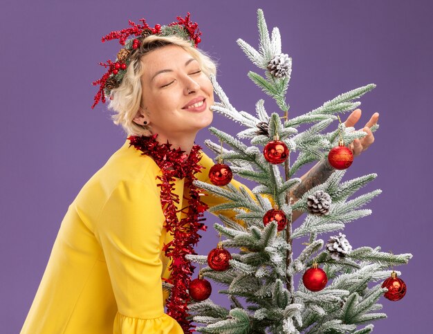 Lachende jonge blonde vrouw hoofd kerstkrans en klatergoud slinger om nek dragen staande in profiel te bekijken in de buurt van versierde kerstboom knuffelen met gesloten ogen geïsoleerd op paarse achtergrond