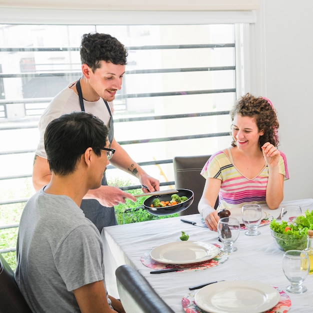 Lachende jonge man eten serveren aan zijn vriend aan tafel zitten