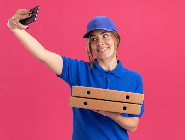 Lachende jonge mooie levering meisje in uniform houdt pizzadozen en kijkt naar de telefoon op roze
