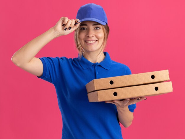 Lachende jonge mooie levering meisje in uniform legt de hand op de dop en houdt pizzadozen op roze