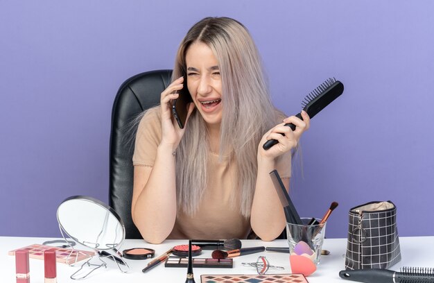 Lachende jonge, mooie meid met een beugel zit aan tafel met make-uptools spreekt op een telefoon met een kam geïsoleerd op een blauwe muur