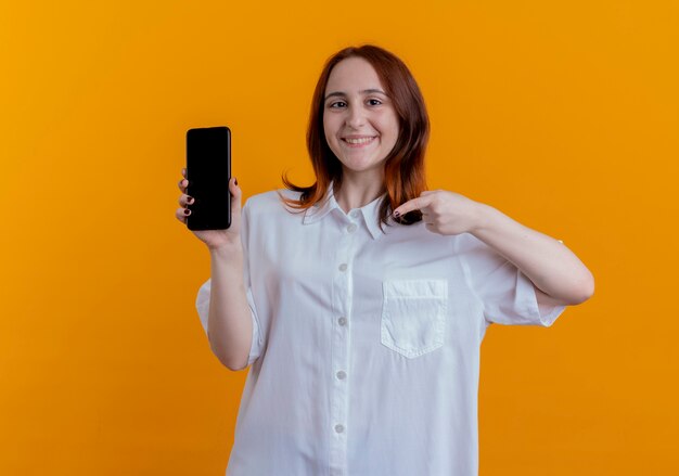 lachende jonge roodharige vrouw met en punten op telefoon geïsoleerd op geel