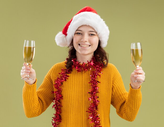 Lachende jonge slavische meisje met kerstmuts en met slinger om nek met glazen champagne geïsoleerd op olijfgroene achtergrond met kopie ruimte