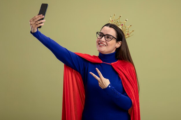 Lachende jonge superheld meisje bril en kroon tonen vredesgebaar neem een selfie geïsoleerd op olijfgroene achtergrond