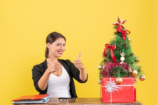 Lachende jonge vrouw in pak ok gebaar maken in de buurt van versierde kerstboom op kantoor op geel