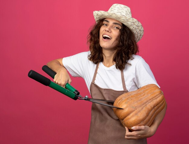 Lachende jonge vrouw tuinman in uniform dragen tuinieren hoed snijden pompoen met tondeuse geïsoleerd op roze